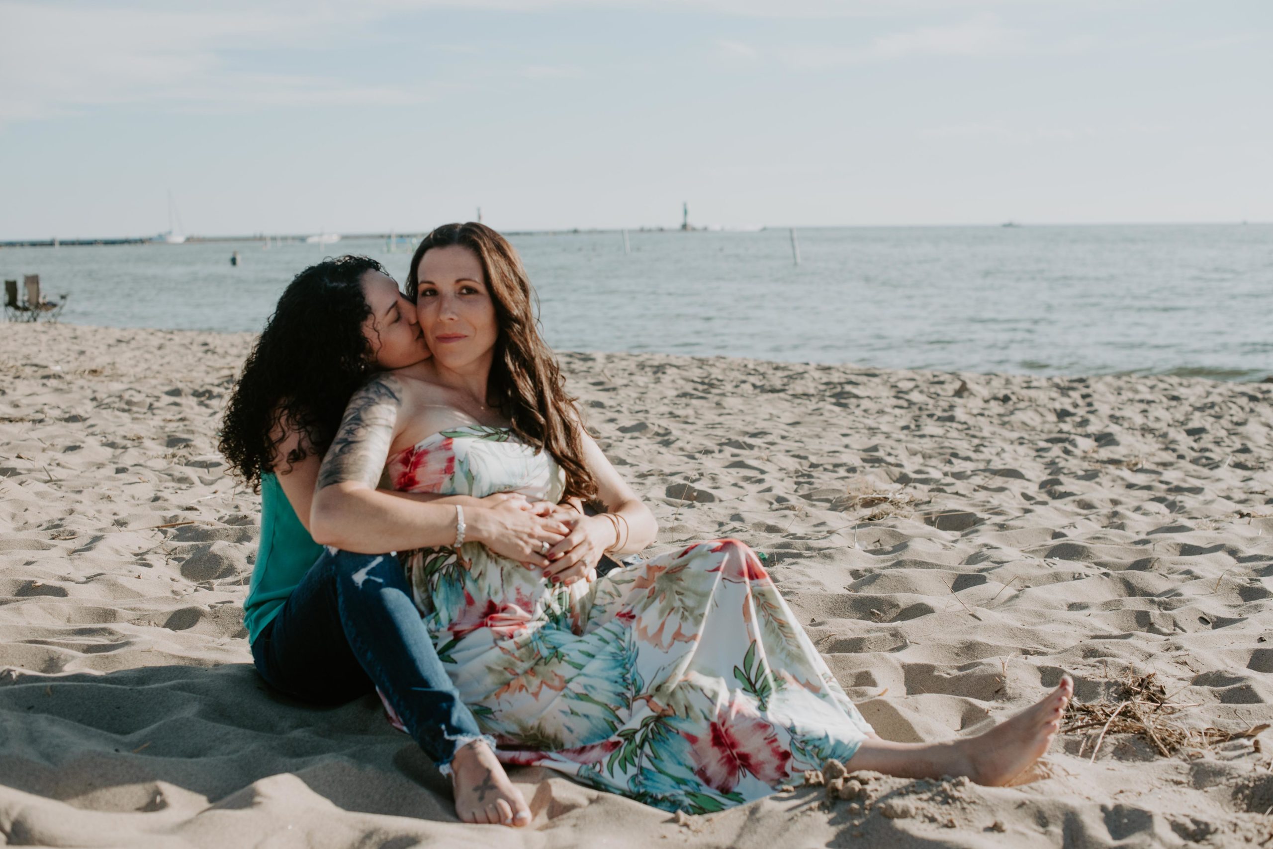same sex maternity photos at holland state park on the beach. Two moms one in a flowy floral print maternity dress and the other in a turquoise top. Lesbian moms LGTBQ maternity photographer in Grand Rapids Michigan. 