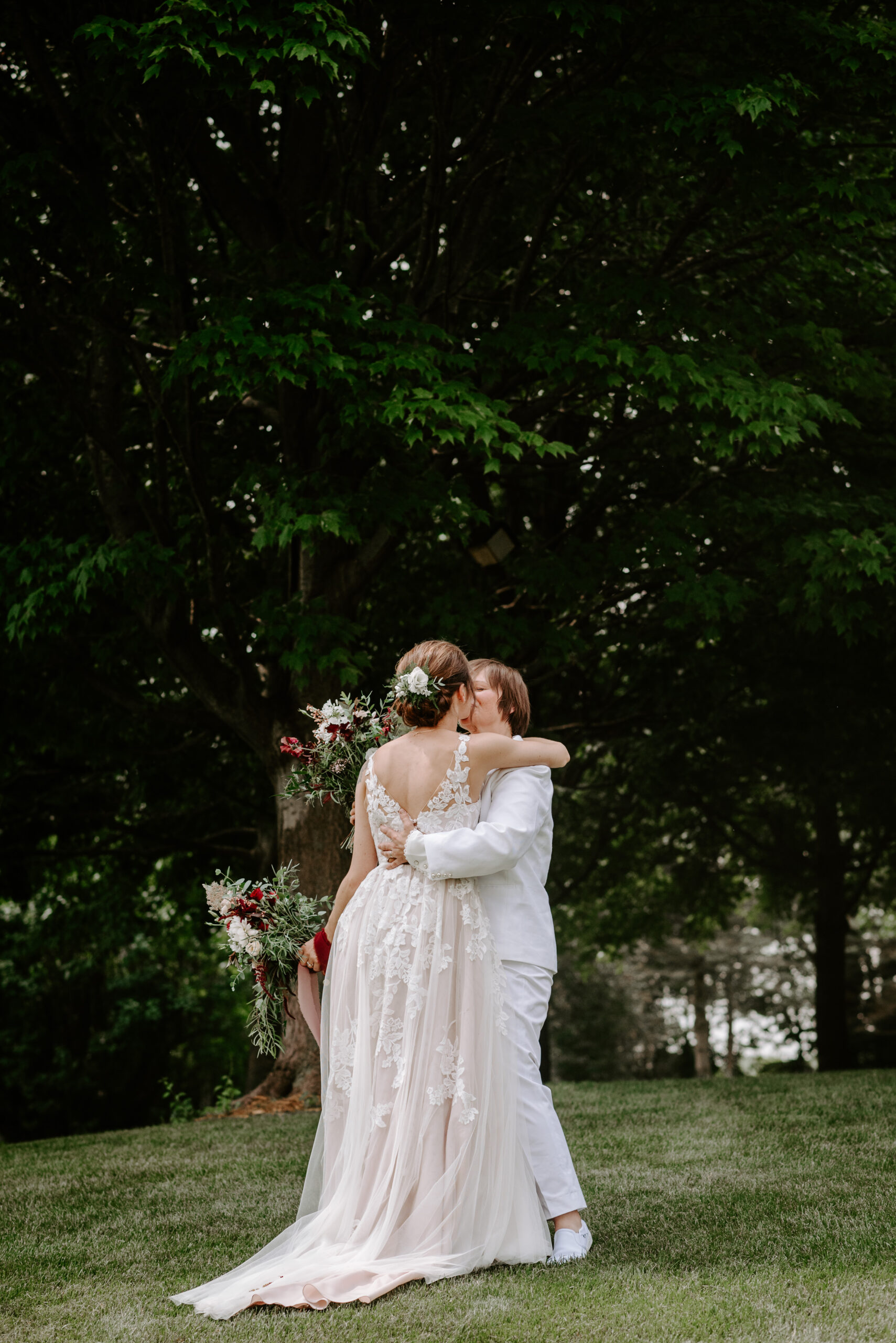 Two brides embracing after their wedding ceremony both wearing white one wearing J.Crew suit one wearing white floral down both holding wedding florals to brides kissing backyard we're winning photographed by Michigan queer wedding photographer lLiv Lyszyk photography based in Grand Rapids
