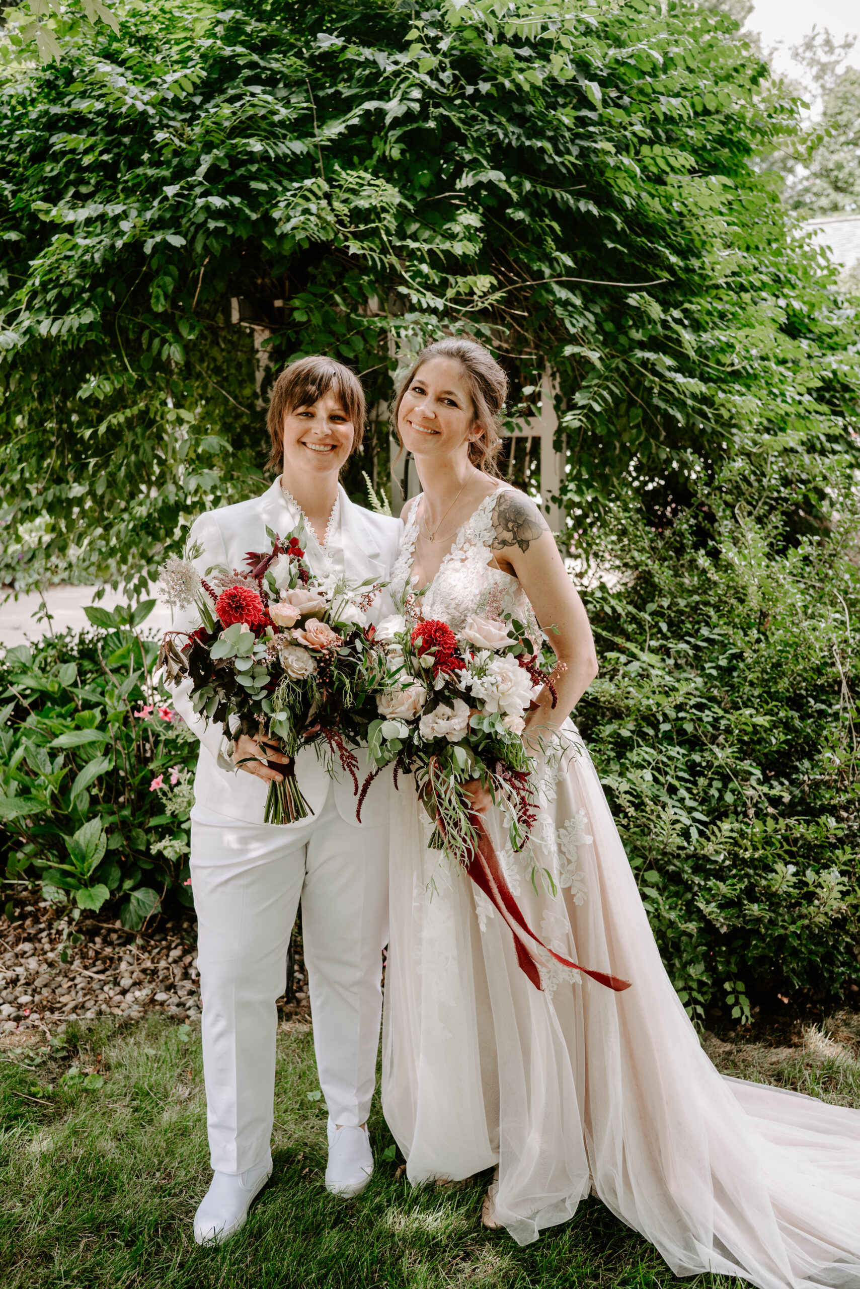 Two brides looking at the camera both in white both holding red and white and pink bouquets LGBTQ backyard wedding summer wedding in Michigan backyard Michigan wedding Lawton Michigan wedding photography by Liv Lyszyk photography who is an LGBTQ plus wedding photographer based in Grand Rapids Michigan