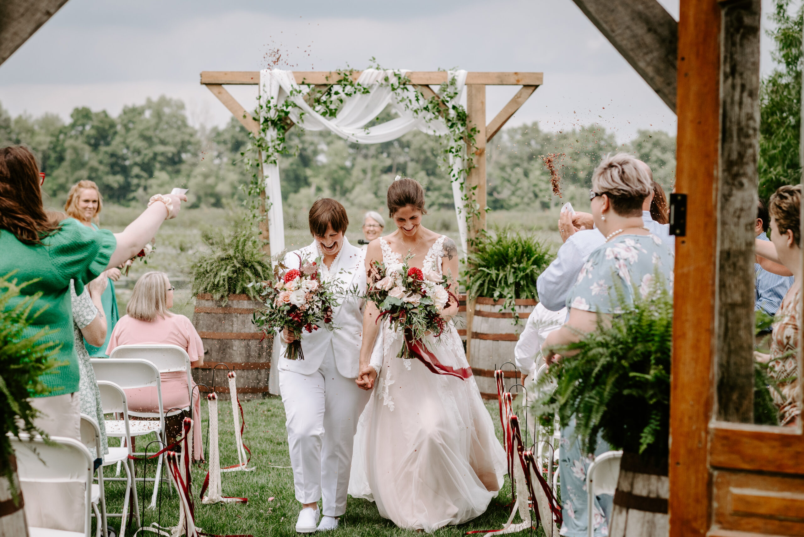 Two brides exiting the ceremony in their LGBTQ plus backyard wedding in Lawton Michigan. One bride wearing white suit from J.Crew other bride wearing white dress both holding wedding florals photographed by queer wedding photographer Liv Lyszyk photography from Grand Rapids Michigan