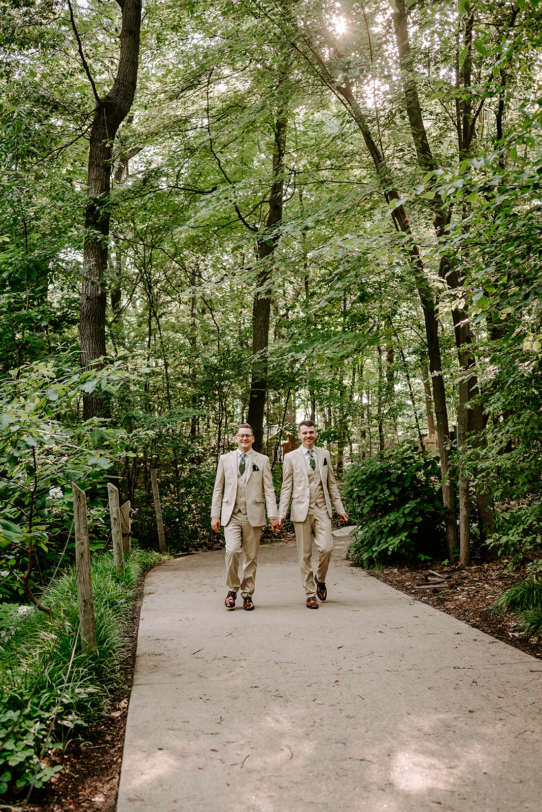 lgbt boho neutral wedding at john ball zoo in grand rapids michigan two grooms in tan suits with green accents walking down pathway with trees
