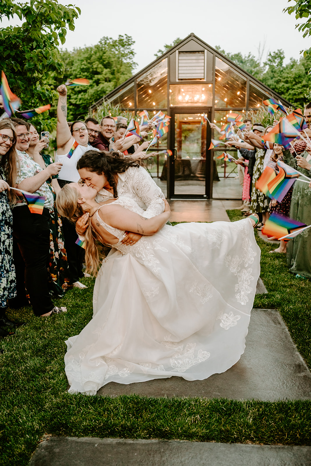 michigan spring DIY lesbian wedding at black sheep shelter exit photo of two brides guests waving rainbow pride flags at sunset