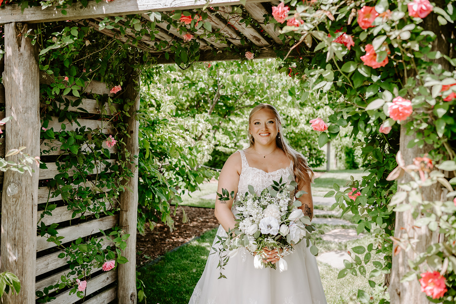 michigan spring DIY lesbian wedding at black sheep shelter blonde bride with white and green florals smiling at camera under floral arch