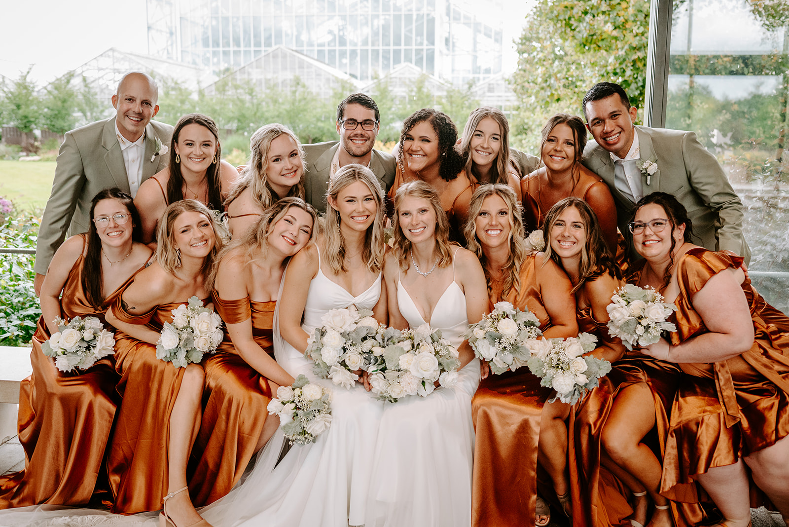 two brides getting married at Meijer gardens at a Grand Rapids LGBT Garden wedding inside the atrium with their entire wedding party in burnt orange silk gowns and gray suits