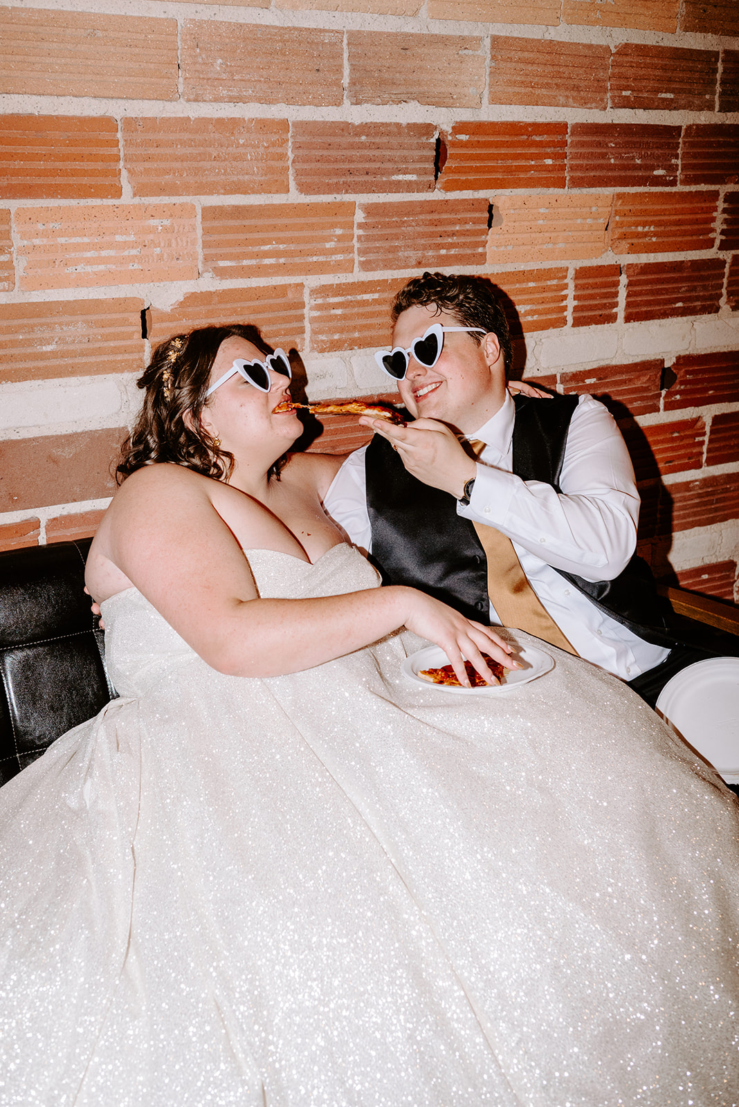 bride and groom sitting on a couch at the end of their wedding night with sunglasses on sharing a piece of pizza