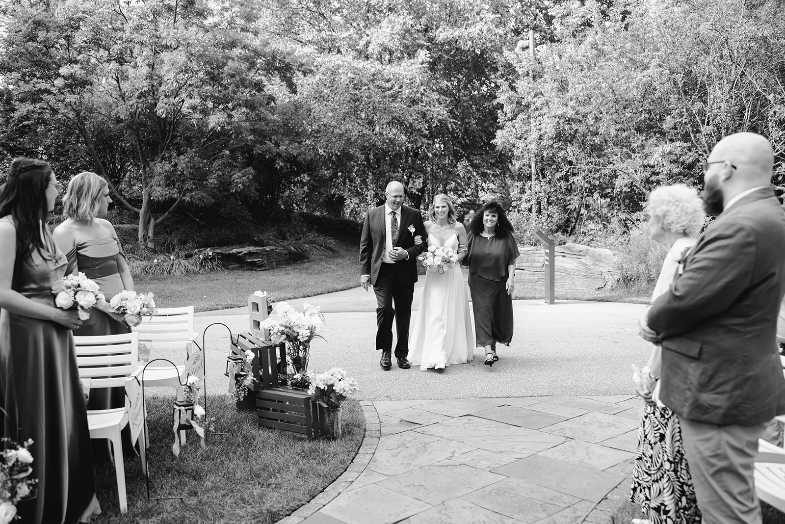 two brides getting married at Meijer gardens at a Grand Rapids LGBT Garden wedding with one bride walking down the aisle with her parents in a black and white image